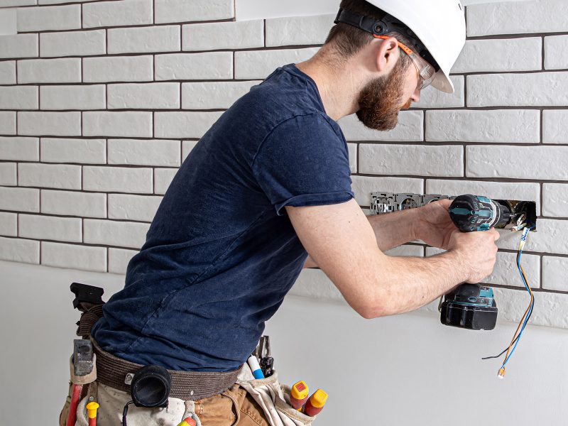 Electrician Builder at work, installation of lamps at height. Professional in overalls with a drill. On the background of the repair site. The concept of working as a professional.